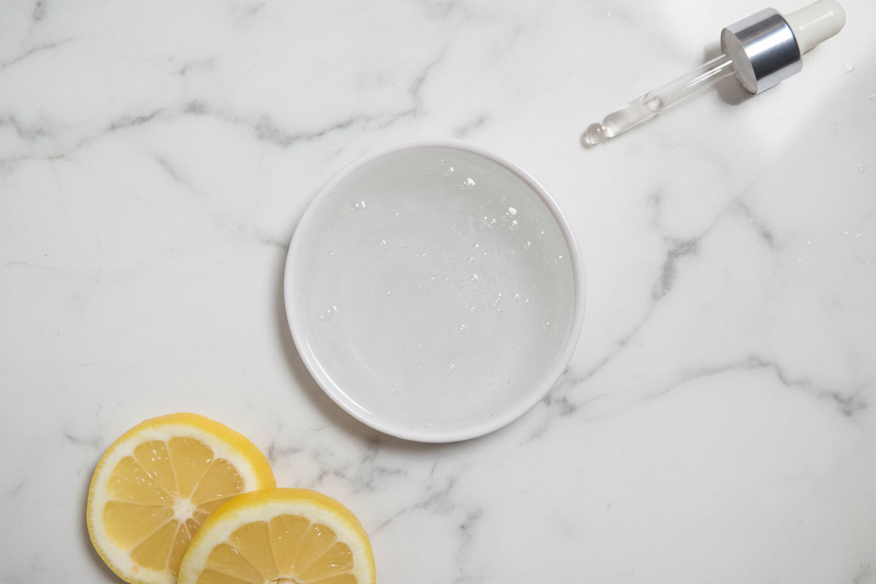 Round small bowl of gel facial cleanser on a whitegrey marble background with lemon slices and a dropper.