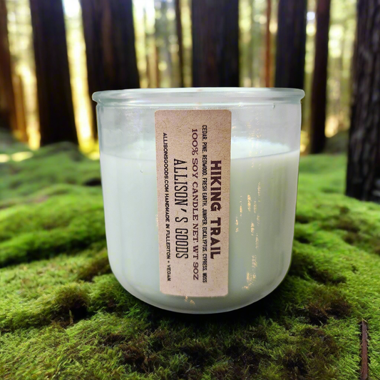 A white soy wax candle in a clear glass jar, placed on a bed of green moss, with a label that reads 'Hiking Trail Soy Candle - California Collection'.