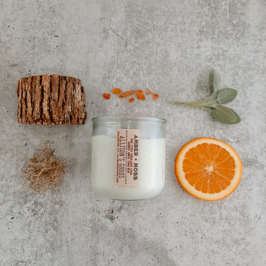 flatlay of vegan candle with amber stones, orange, moss, wood, and sage leaves